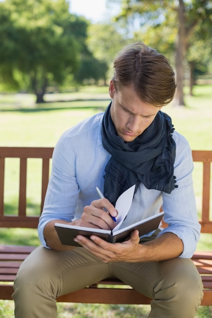 Élégant jeune homme écrivant dans son bloc-notes sur un banc de parc