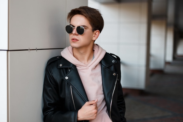 Élégant jeune homme hipster avec une coiffure à la mode dans des lunettes noires élégantes dans une veste en cuir noir dans un sweat-shirt rose se dresse près d'un mur blanc. Beau mec américain. La mode masculine.