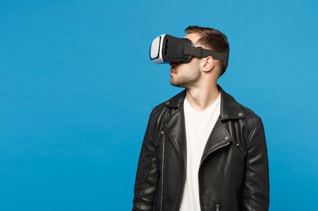 Élégant jeune homme mal rasé en t-shirt blanc veste noire regardant dans un casque, réalité virtuelle vr isolé sur fond de mur bleu portrait en studio. Concept de mode de vie des émotions des gens. Maquette de l'espace de copie.