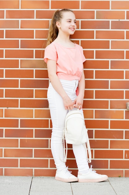 Élégant de la tête aux pieds Petit mannequin au look élégant Petit enfant mignon avec de longs cheveux blonds dans des vêtements de mode élégants Adorable fille élégante sur un mur de briques rouges