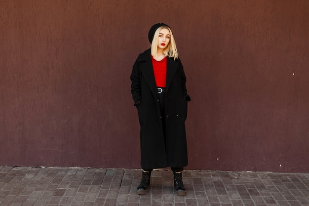 Élégante belle femme dans un manteau noir à la mode avec des bottes posant près d'un mur