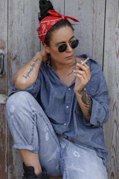 Élégante belle femme hipster américaine en denim vêtements à la mode avec un bandana rouge et des lunettes de soleil fumant une cigarette et assis près d'un mur en bois, tatouage