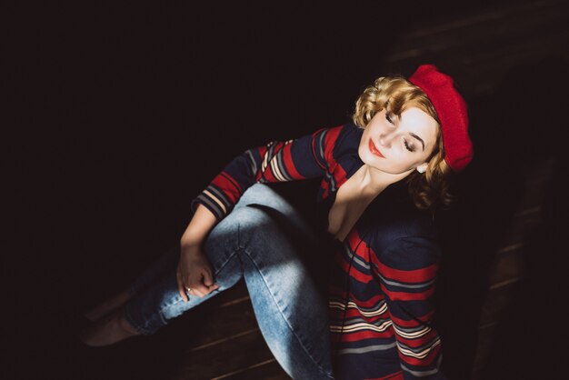 Élégante belle fille blonde dans un béret rouge est assis sur un plancher en bois sombre au soleil.