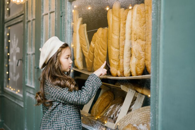 Élégante, belle fille de six ans dans un béret blanc et un manteau à carreaux vert pointe vers une baguette dans une fenêtre de boulangerie