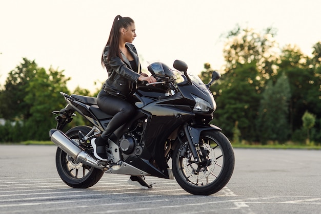 Élégante belle fille en veste et pantalon en cuir noir sur un parking extérieur se trouve sur le sport