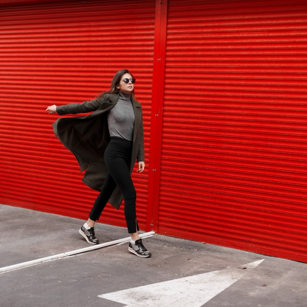 Élégante belle jeune femme avec des lunettes de soleil dans un manteau de mode se promène dans la rue près des portes en métal rouge.