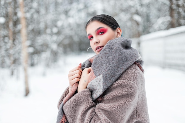 Élégante belle jeune femme avec un maquillage rose aux couleurs vives avec des ombres à paupières dans un vêtement d'hiver à la mode avec une écharpe et un manteau de fourrure dans la forêt avec de la neige