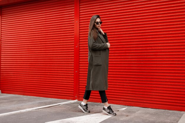 Élégante belle jeune femme séduisante avec des lunettes de soleil dans un manteau à la mode sur un mur rouge marchant dans la rue. Mode de rue