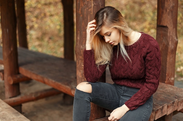 Élégante belle jeune fille dans un pull tricoté à la mode se trouve et repose près d'une maison en bois sur la nature