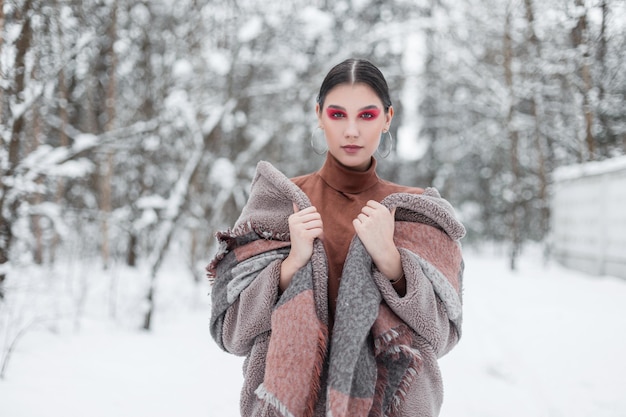 Élégante belle jeune fille hipster avec un maquillage rose vif dans des vêtements à la mode d'hiver posant dans la forêt de neige