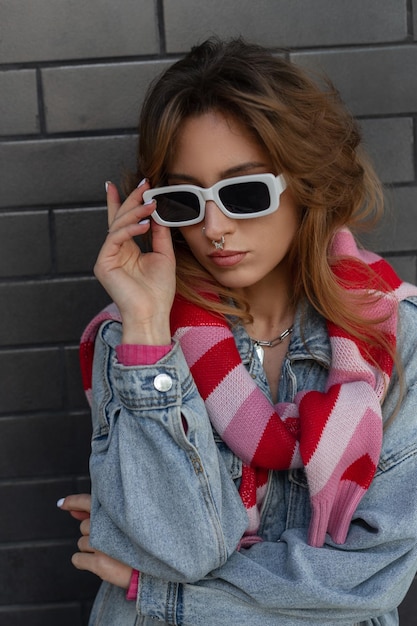 Élégante belle jeune fille modèle rousse avec un piercing au nez dans une tenue de rue à la mode avec une veste en jean et un sweat-shirt rose porte des lunettes de soleil de mode blanches fraîches près d'un mur de briques noires