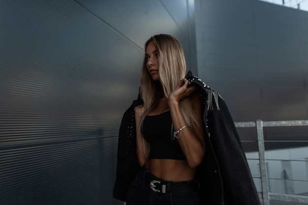 Élégante belle jeune fille modèle en veste de jeans à la mode noire, short en jean et débardeur posant près d'un mur moderne en métal dans la rue. Tenue de style féminin urbain