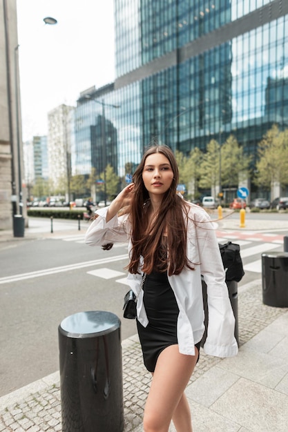 Élégante belle jeune fille de rue dans des vêtements de mode avec une chemise blanche et une robe noire se promène dans la ville