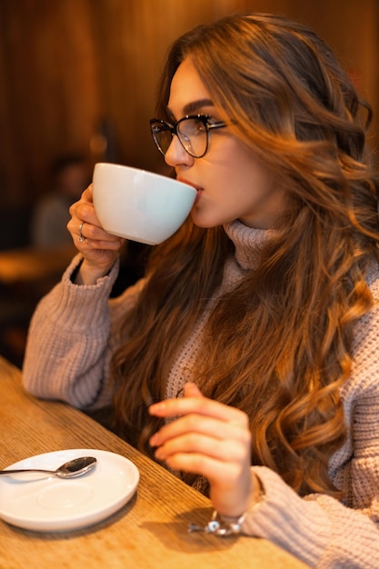 Élégante belle jeune jolie fille à la mode des vêtements chauds avec un pull vintage tricoté est assis dans un café et boit du café