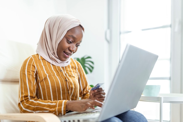 Élégante femme musulmane séduisante utilisant un ordinateur portable mobile à la recherche d'informations sur les achats en ligne dans le salon à la maison. Portrait d'une femme heureuse achetant un produit via des achats en ligne. Payer par carte de crédit