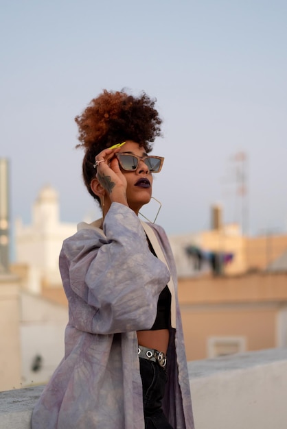 Élégante femme noire aux cheveux bouclés et lunettes de soleil portant une robe lilas tout en se reposant sur une terrasse contre un ciel bleu sans nuages à Cadix Espagne