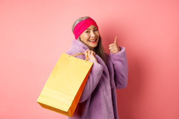 Élégante femme senior asiatique faire du shopping, porter un sac en papier sur l'épaule et montrant le pouce vers le haut, recommandant des remises en magasin sur le rose.