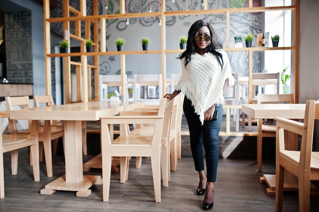 Élégante fille afro-américaine à lunettes de soleil posée au café moderne