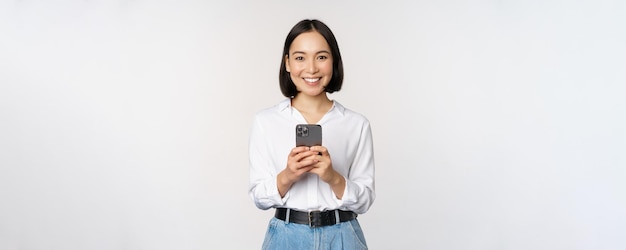 Élégante fille asiatique moderne à l'aide d'une application de téléphonie mobile discutant sur un téléphone portable et souriant debout en blouse blanche sur fond de studio