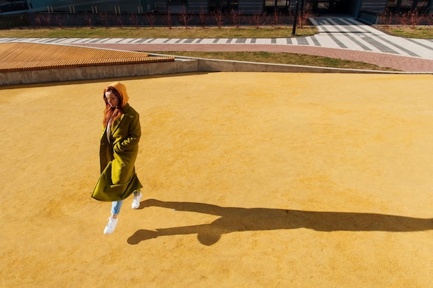 Élégante fille aux cheveux roux en long manteau vert et lunettes de soleil marchant dans la rue au printemps ensoleillé Longue ombre au sol jaune d'ici