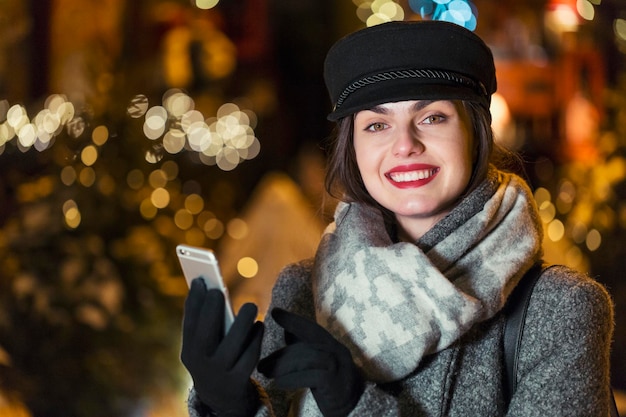 Élégante fille brune souriante à l'aide d'un smartphone devant le mur de guirlandes dans la soirée de Noël de la ville