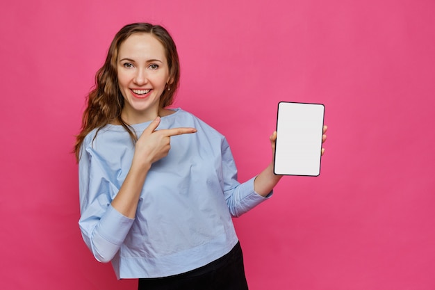 Élégante fille caucasienne dans un t-shirt bleu pâle tient une tablette à la main et pointe un doigt dessus sur un fond rose.