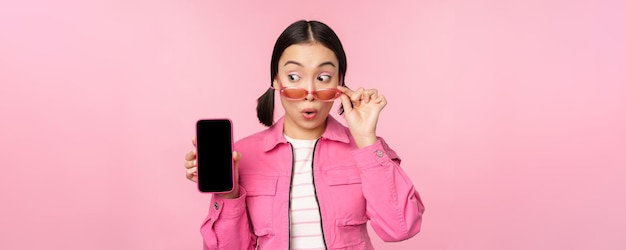 Élégante fille coréenne jeune femme à lunettes de soleil montrant l'interface de l'application de téléphone mobile à écran de smartphone ou le site Web debout sur fond rose