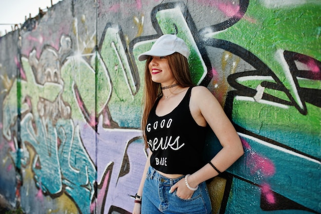 Élégante fille hipster décontractée en casquette et jeans porter contre un grand mur de graffitis