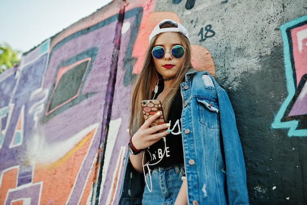 Élégante fille hipster décontractée en casquette lunettes de soleil et jeans porter écouter de la musique à partir d'écouteurs de téléphone portable contre un grand mur de graffitis