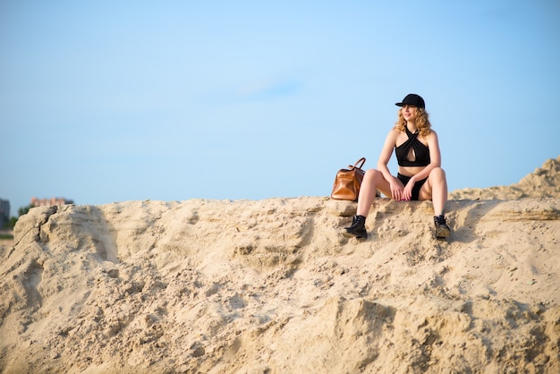 Élégante jeune et belle femme dans un sous-vêtement noir portant une casquette et des bottes en cuir avec un sac en cuir se trouve sur une carrière de sable par une chaude journée d'été ensoleillée.