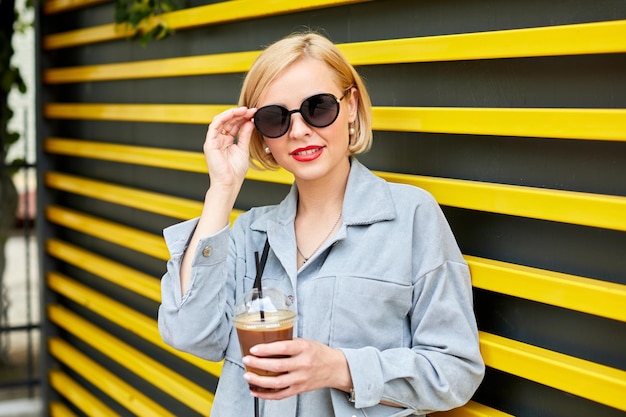 Élégante jeune blonde à lunettes de soleil se promène dans la ville et boit une boisson fraîche