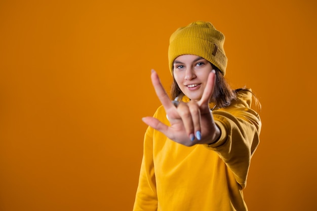 Élégante jeune brune gaie dans un sweat à capuche jaune et des chapeaux montre un geste cool deux doigts sur un...