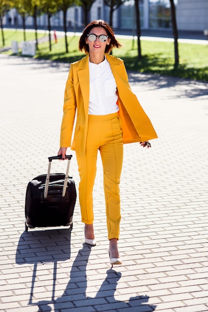 Élégante jeune femme d'affaires en costume jaune élégant se précipite pour une réunion d'affaires, parle sur smartphone et tire une valise. Femme d'affaires attrayant partant en voyage d'affaires en tirant sa valise.