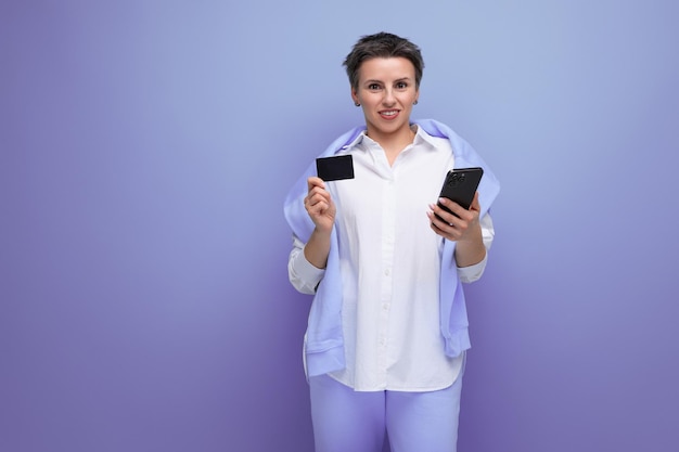 Élégante jeune femme brune avec une coupe de cheveux courte utilise un smartphone et une carte pour payer ses achats