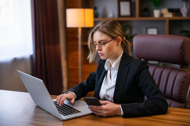 Élégante jeune femme entrepreneur assise à un bureau dans son bureau à domicile travaillant en ligne avec un ordinateur portable