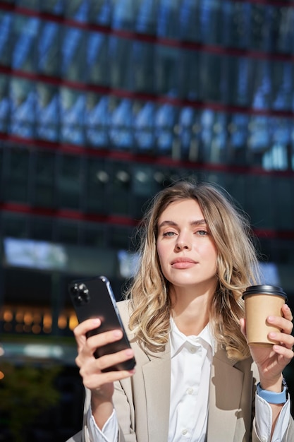 Élégante jeune femme d'entreprise debout dans la rue boit du café et tient un téléphone portable