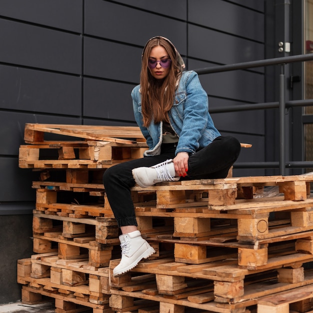 Élégante jeune femme hipster assez cool dans des vêtements de jeans à la mode dans des lunettes à la mode violettes en bottes blanches en cuir assis sur des palettes en bois près du mur dans la rue. Belle fille urbaine. Style décontracté