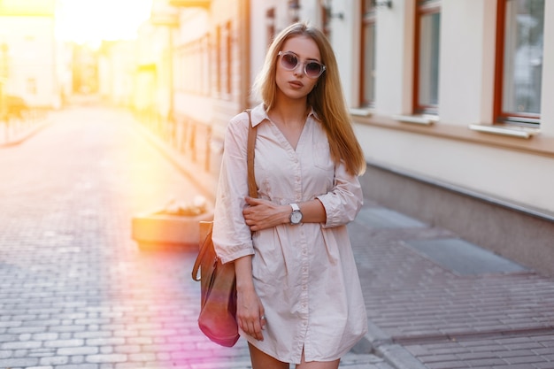 Élégante jeune femme hipster dans des lunettes de soleil à la mode dans une robe à la mode d'été en baskets blanches avec un sac à main en cuir élégant se tient debout dans la ville par une chaude journée ensoleillée au coucher du soleil.