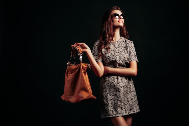 Élégante jeune femme à la mode en lunettes de soleil portant une robe imprimée serpent tout en tenant un sac à main marron