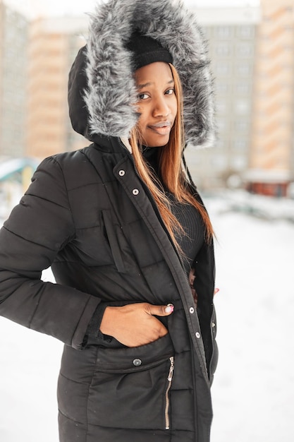 Élégante jeune femme noire dans une veste à la mode avec une capuche se promène dans la rue avec de la neige