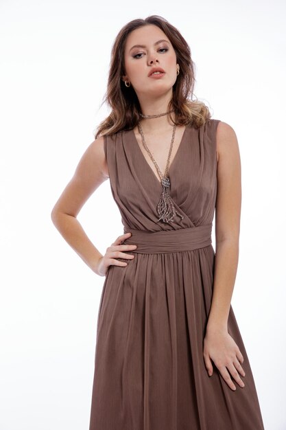 Élégante jeune femme en robe marron beige avec un collier décolleté posant sur fond blanc