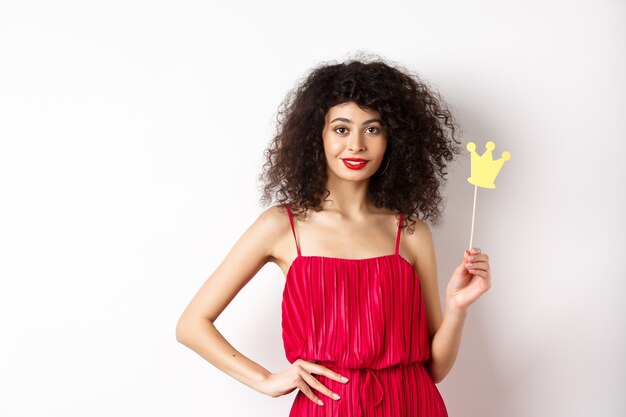 Élégante jeune femme en robe rouge, se sentant confiante et impertinente, tenant la couronne et souriant, debout sur fond blanc.