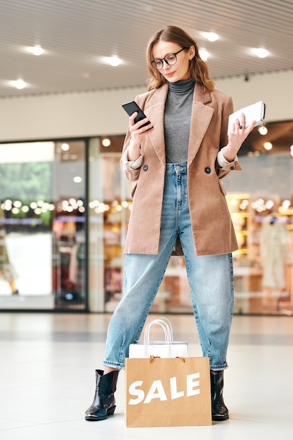 Élégante jeune femme trouver des rabais dans les magasins avec application pour smartphone tout en faisant du shopping dans le centre commercial