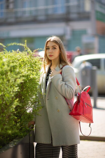 Élégante jeune jolie fille dans un manteau gris regarde la caméra et sourit légèrement dans le contexte de la ville. La jeune fille se tient près d'un buisson vert.