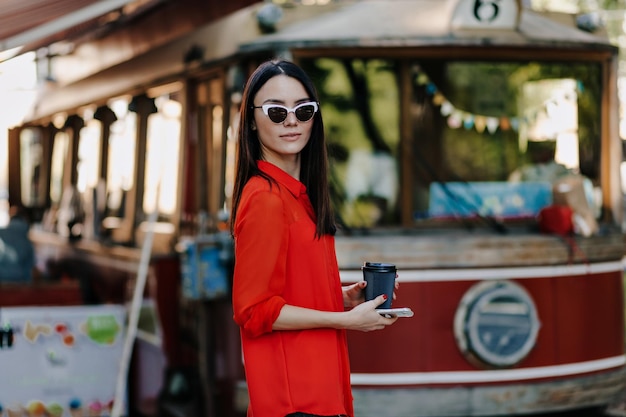 Élégante jolie femme aux cheveux raides foncés portant un chemisier rouge à lunettes de soleil tient un café pour aller marcher dans la ville par une chaude journée de printemps