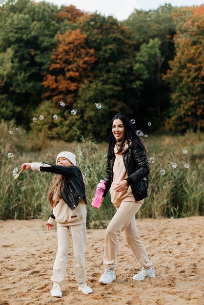 Élégante maman et sa fille en vestes de cuir soufflent des bulles dans un parc d'automne