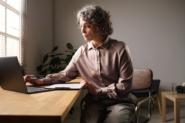 Élégante psychologue mature assise sur le lieu de travail devant un ordinateur portable