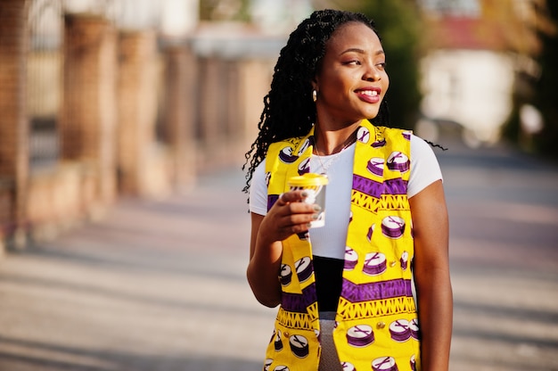 Élégantes femmes afro-américaines en veste jaune posés dans la rue avec une boisson chaude dans une tasse en papier jetable.