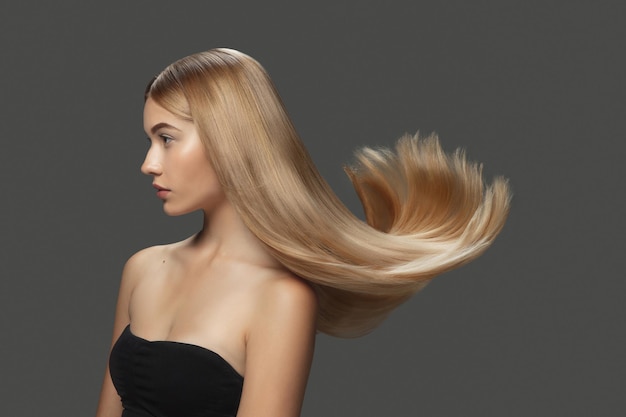 Léger. Beau modèle avec de longs cheveux blonds lisses et volants sur fond de studio gris foncé. Jeune mannequin caucasien avec une peau bien entretenue et des cheveux soufflant dans l'air. Concept de soins de salon, beauté, mode.