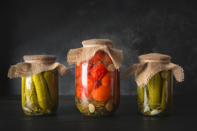 Légumes en conserve concombre et tomate dans des bocaux en verre sur planche de bois noir Vue latérale Récolte maison et préparations d'automne